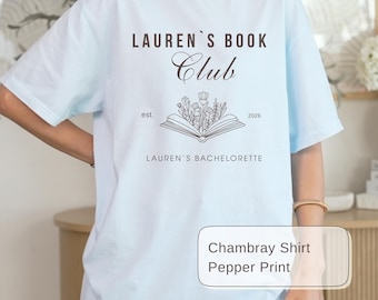 May include: Light blue t-shirt with the text "LAUREN'S BOOK Club" above an open book illustration with flowers. Below the illustration is the text "LAUREN'S BACHELORETTE". The shirt also has the text "est. 2026".