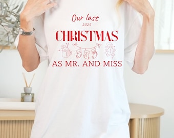 Op de afbeelding: Wit T-shirt met rode tekst: "Our last CHRISTMAS 2025 AS MR. AND MISS." De tekst wordt vergezeld door feestelijke illustraties van een mok, kousen en een peperkoekmannetje. Het shirt heeft een relaxte, oversized pasvorm.