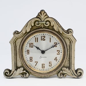 May include: A mantel clock with a metallic, textured frame featuring ornate scrollwork and a pointed top. The circular clock face has a white background, black Arabic numerals, black hands, and the word Sessions printed in black script.