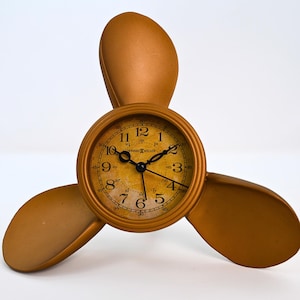 May include: A bronze-colored clock in the shape of a propeller. The clock face has black hands and numerals. The clock is set against a white background.