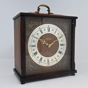 May include: A rectangular wooden mantel clock with a dark brown finish and a brass handle on top. The clock face features Roman numerals, gold-colored ornate hands, and the text Elgin. The corners of the glass front display intricate gold filigree patterns.