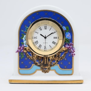 Puede incluir: Un reloj de cuarzo de cerámica blanca con bisel dorado y números romanos. La esfera del reloj está rodeada por un diseño decorativo azul y dorado con detalles florales. La base tiene un borde dorado.