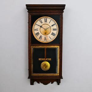 May include: A rectangular wall clock with a dark brown wooden frame. The upper section features a circular white face with black Roman numerals and hands. The lower section has a black glass panel with the word REGULATOR and a gold pendulum.