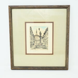 May include: Framed artwork featuring a detailed pen and ink drawing of a European city square. The artwork is set within a brown wooden frame and a cream-colored mat. The drawing depicts buildings, a clock tower, and a fountain.
