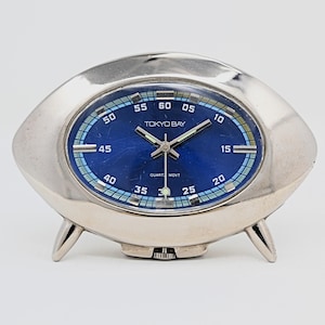 May include: A silver-toned Tokyo Bay quartz movement clock with a blue face. The clock has a unique, rounded shape with three small feet. The face displays the time with white numerals and hands. The clock's face has a blue and light blue design.