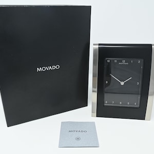 Könnte beinhalten: Eine schwarze und silberne Movado-Schreibtischuhr mit quadratischem Zifferblatt. Die Uhr hat ein schwarzes Zifferblatt mit silbernen Zeigern und Stundenmarkierungen. Die Uhr befindet sich in der Originalverpackung mit dem Movado-Logo auf der Schachtel und einer kleinen Karte.