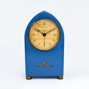 May include: A blue mantel clock with a pointed arch top and a gold-colored circular face. The clock features black hands, gold-colored numerals, and a grid-patterned background on the dial. A small triangular decorative cutout is located below the face.