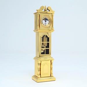 Puede incluir: Un reloj de pie dorado con una esfera blanca y una parte superior decorativa. El reloj tiene un cuerpo rectangular con una sección cubierta de vidrio que revela el mecanismo interno. Es un artículo decorativo clásico.