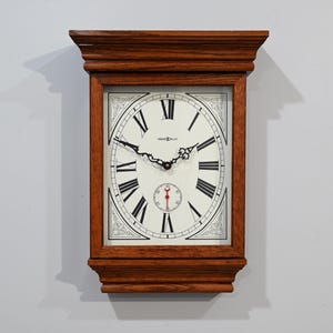 Pode incluir: Um relógio de parede retangular de madeira com um mostrador claro e algarismos romanos pretos. O relógio tem um pequeno mostrador de segundos e ponteiros decorativos. A moldura de madeira é de cor castanho escuro.