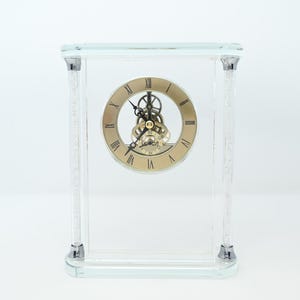 May include: Clear glass clock with a gold-tone face and Roman numerals. The clock features a visible mechanical movement and is encased in a rectangular glass frame with crystal accents. A decorative timepiece for a desk or shelf.