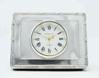 Può includere: Un orologio da tavolo rettangolare color argento con una cornice metallica lucida caratterizzata da un motivo a bordo in rilievo. Il quadrante bianco mostra numeri romani neri, lancette dorate e la scritta Kitney & Co London al centro.
