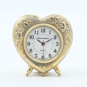 May include: A heart-shaped quartz clock with a gold-colored metal frame. The clock face is white with black numerals and hands, and the frame features cherub and heart designs. The clock is marked "MILAN" and "JAPAN MOVT".
