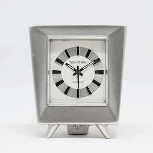 May include: A silver-toned Tokyo Bay quartz clock with a square face and a unique, angled frame. The clock face is white with black and silver radial stripes and the words "TOKYO BAY" and "QUARTZ". The clock has three small feet.