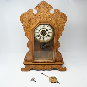 Puede incluir: Un reloj antiguo de madera con intrincadas tallas y una esfera de cristal. La esfera del reloj es blanca con números romanos negros. Se incluye una llave y un péndulo. El reloj mide aproximadamente 46 cm de alto.