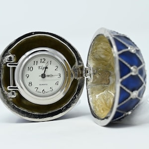 May include: An open, decorative egg-shaped clock with a silver-tone exterior and a blue and silver patterned design on the lid. The clock face is white with black numerals and the brand name "Elgin".