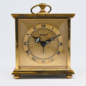May include: A gold-toned Jerger clock with a square face and Roman numeral hour markers. The clock features a decorative face with a circular dial, black hands, and a small handle on top. The clock is supported by four small, rounded feet.