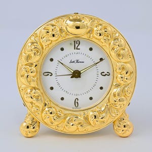 May include: A round gold-colored table clock with an ornate, embossed scrollwork frame. The white clock face features black Arabic numerals at the 3, 6, 9, and 12 positions, black hands, and the text Seth Thomas and Germany printed on the dial.