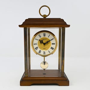 May include: A rectangular mantel clock with a dark wood base and top, featuring a brass handle on the summit. The clock has a glass-enclosed body, a circular face with black Roman numerals, gold-toned center, and a swinging brass pendulum.