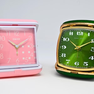 May include: Two vintage travel clocks. One is pink with a white frame and a pink clock face. The other is green with a gold frame. Both clocks have white numbers and gold hands. The pink clock has the brand name "EQUITY" on the clock face.