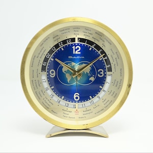 May include: A vintage Bulova world time clock with a gold-tone bezel and a blue face featuring a world map. The clock displays multiple time zones around the perimeter. The clock has gold hands and a silver base.