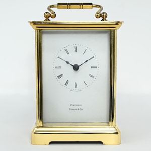 May include: A gold-toned brass carriage clock with a rectangular frame and a top handle. The white clock face features black Roman numerals, thin black hands, and the text "PORTFOLIO" and "TIFFANY & CO." printed in black on the lower section.