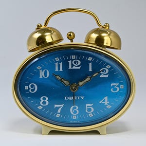 May include: An oval-shaped alarm clock with a metallic gold frame and two bells on top. The clock face is a vibrant blue with white Arabic numerals from 1 to 12, ornate hands, and the brand name Equity printed in white text in the center.