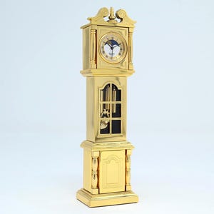 May include: A miniature gold-toned grandfather clock featuring a circular clock face with Roman numerals, black hands, and a moon phase dial. The central section displays a visible pendulum and weights behind a glass panel, set on a base.