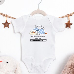 May include: White baby onesie with a humorous graphic of a sleeping duck wearing a nightcap and the text "Please hold, I'm mentally buffering to be silly." The onesie is hanging on a white hanger.