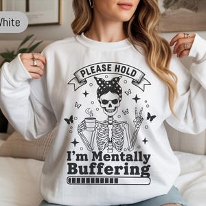 May include: White sweatshirt with a black and white skeleton graphic. The skeleton is wearing a polka dot headband and holding a coffee cup. Text on the sweatshirt reads "PLEASE HOLD" and "I'm Mentally Buffering."