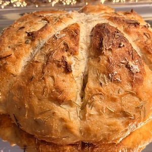 May include: A close-up shot of a round loaf of bread, golden brown with a crusty exterior. The bread is decorated with rosemary sprigs. The bread is sitting on a reflective surface.