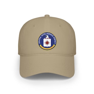 Puede incluir: Gorra de béisbol beige con el logotipo de la Agencia Central de Inteligencia (CIA). El logotipo es un diseño circular con las palabras "CENTRAL INTELLIGENCE AGENCY" y "UNITED STATES OF AMERICA" que rodean un emblema central de un águila y una estrella.