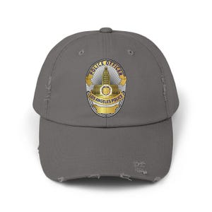 May include: A grey distressed baseball cap displaying the Los Angeles Police Officer badge. The badge is gold and yellow with the words "POLICE OFFICER" and "LOS ANGELES POLICE" in black text. The cap has a curved peak and a distressed appearance.