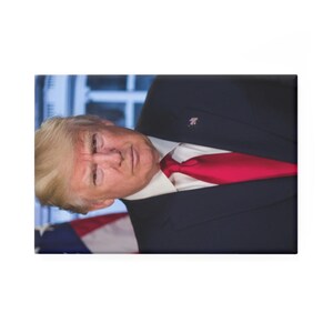 Op de afbeelding: Een portret van een man in een donker pak, wit overhemd en een felrode stropdas. De man heeft licht haar en kijkt naar de kijker. Een Amerikaanse vlag is zichtbaar op de achtergrond.