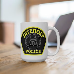 May include: White ceramic mug featuring the Detroit Police logo. The logo is a black and yellow emblem with the words "DETROIT" and "POLICE" and the text "AUTOMOTIVE CAPITAL OF THE WORLD". The mug has a large handle.