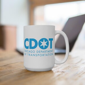 May include: White ceramic mug with the text "CDOT CHICAGO DEPARTMENT OF TRANSPORTATION" in blue. The mug has a large handle and is sitting on a wooden surface. A laptop is visible in the background.