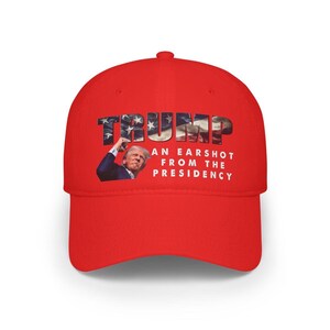 May include: A red baseball cap with the word "TRUMP" in a patriotic font. The cap features an image of a person and the text "AN EARSHOT FROM THE PRESIDENCY".