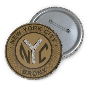 May include: A round button with a brown face featuring the text "NEW YORK CITY BRONX" around the edge. The center has an "NYC" design filled with a textured gray material. The back is silver with a pin.