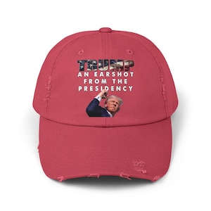 May include: A distressed red baseball cap with the word "TRUMP" in a patriotic font. The text "AN EARSHOT FROM THE PRESIDENCY" is below the word "TRUMP". An image of a man is below the text.