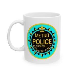 May include: White ceramic mug featuring the Metro Police Nashville Davidson County logo. The logo is a circular design with a teal border, black center, and yellow accents. The text "METRO POLICE" and "NASHVILLE DAVIDSON COUNTY" are displayed.