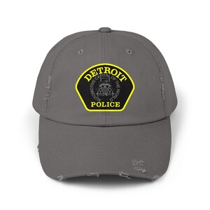 May include: A distressed gray baseball cap featuring the Detroit Police Department emblem. The emblem is a black shield with a yellow border, and the text "DETROIT AUTOMOTIVE CAPITAL OF THE WORLD POLICE" is printed on it.