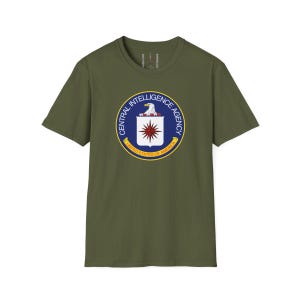 Puede incluir: Camiseta verde oliva con el emblema de la Central Intelligence Agency (CIA). El emblema es un diseño circular con un águila blanca, una estrella roja y blanca, y el texto "CENTRAL INTELLIGENCE AGENCY" y "UNITED STATES OF AMERICA".