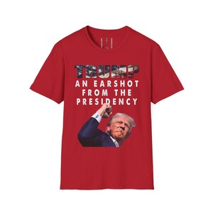 May include: Red t-shirt with the word "TRUMP" in a patriotic font, and the phrase "AN EARSHOT FROM THE PRESIDENCY." Below the text is an image of a man in a suit.