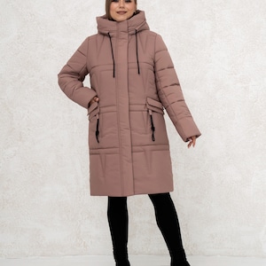 May include: A long, dusty rose-colored puffer coat with a hood and black drawstrings. The coat has two front pockets and a zip closure. The person is wearing black leggings and black boots.
