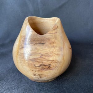 May include: A small, rounded wooden vessel with a smooth, polished finish. The object features a light tan colour with prominent natural wood grain patterns, dark brown streaks, and a wide, open top with a curved, dip-shaped rim design.