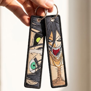 Puede incluir: Dos llaveros bordados con caras de personajes de anime. Uno muestra un ojo verde y un ojo amarillo, el otro un personaje con la boca abierta y dientes afilados. Ambos tienen bordes negros y anillos plateados. Aproximadamente 10 cm de largo.