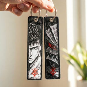 Peut inclure: Deux porte-clés rectangulaires noirs avec des anneaux argentés. Chaque porte-clés présente une illustration détaillée en noir et blanc d'un personnage de manga. Un symbole rouge est en bas de chaque porte-clés.