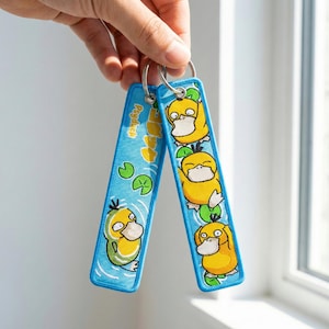 Puede incluir: Dos llaveros azules con un diseño de dibujos animados de una criatura amarilla con forma de pato. Los llaveros tienen un fondo azul claro con la palabra "Psyduck" y la imagen de la criatura. Los llaveros están unidos por un anillo de metal plateado.