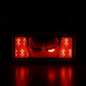May include: A red illuminated display showing a close-up of a character's face with a determined expression. The image is framed by a black border and includes white symbols on the sides. The overall theme is likely related to anime or manga.