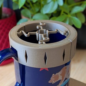 Peut inclure: Une tasse en céramique bleu marine avec une anse, ornée du drapeau de la Californie. Un couvercle beige imprimé en 3D avec des découpes en forme de losange et un motif central est posé sur la tasse. La tasse est sur une surface en bois, avec des plantes en arrière-plan.