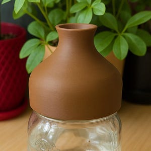 May include: A clear glass jar with a brown lid, filled with water. The jar has a floral design etched into the glass. The lid is a matte brown color. Green plants are visible in the background.